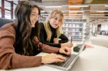 Zwei Frauen sitzen vor einem Laptop und überlegen zusammen, was sie schreiben.
