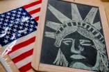 Auf einem Tisch liegen eine Amerika-Flagge und eine Schiefertafel, auf der der Kopf der Freiheitsstatue mit Kreide gezeichnet ist.