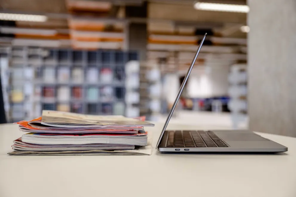Zeitschriften und ein Laptop stehen auf einem Tisch, im Hintergrund sind Bücherregale zu sehen.