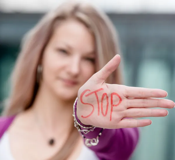 Photo: A woman holds her flat hand up to the camera. The word "Stop" is written in red capital letters on the palm of her hand. The hand is in the foreground, the woman's face is in the background and slightly out of focus.